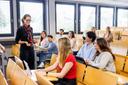 Eine Dozentin steht zwischen den Riehen in einem Hörsaal und erklärt etwas. Mit freundlichem Blick schaut sie eine Studentin an. Die übrigen Studierenden blicken in Richtung der Dozentin.