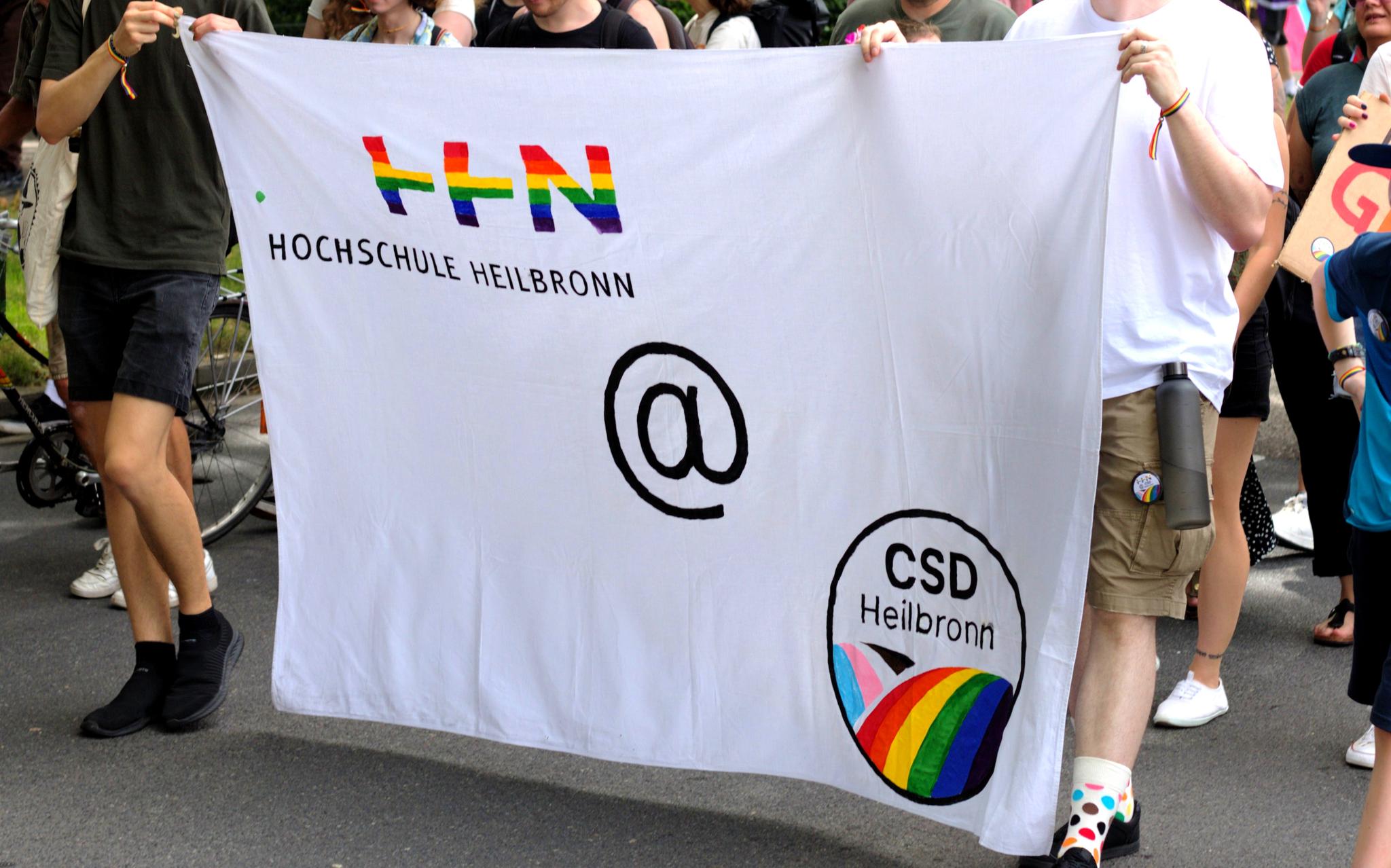 Das Foto zeigt eine Gruppe von Menschen, die am CSD Heilbronn teilnehmen. Sie halten ein großes weißes Banner mit der Aufschrift "Hochschule Heilbronn" und einem Logo, das die Buchstaben "HHN" in Regenbogenfarben zeigt. Unten rechts auf dem Banner befindet sich ein weiteres Logo mit der Aufschrift "CSD Heilbronn" und einem Regenbogenmotiv. Die Teilnehmende tragen lässige Kleidung, einige mit Regenbogenarmbändern, und es ist ein Fahrrad im Hintergrund.