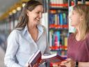 Professorin reicht lachend einer Studentin in der Bibliothek ein Buch.
