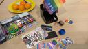 Auf einem hellen Holztisch liegen bunte Filzstifte, Sticker mit Regenbogenmotiven, zwei Rollen Washi-Tape und ein kleiner Organizer mit einer Regenbogenfahne. Im Hintergrund steht ein gelber Teller mit Mandarinen. Die Sticker zeigen verschiedene Motive wie Schriftzüge, Herzen und Hände in Regenbogenfarben. Die Szene wirkt kreativ und einladend.