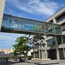 Moderner Hochschulcampus mit zwei mehrstöckigen Gebäuden aus hellem Mauerwerk, verbunden durch eine verglaste Brücke. Auf der Brücke hängt ein Banner mit der Aufschrift „Du bist hier richtig“. Davor ein gepflasterter Platz mit Sitzbänken, Bäumen und einzelnen Personen. Blauer Himmel mit vereinzelten Wolken.