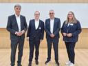 Drei Männer und eine Frau in Businesskleidung vor einem neutralen Hintergrund.
