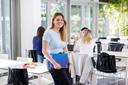 Eine Studentin lehnt an einem Tisch, hält eine Mappe unter dem Arm und blickt lächelnd und offen direkt in die Kamera
