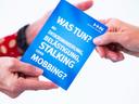 Eine Person übergibt einer anderen einen Flyer mit der Aufschrift "Was tun bei Diskriminierung, Belästigung, Stalking und Mobbing?"