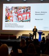 Foto von Robin Afamefuna auf einer Bühne. Im Hintergrund eine Powerpoint-Präsentation, worauf ein Foto von Robin Afamefuna im Fußbaltrikot mit Megafon abgebildet ist. Darunter steht: "Zwischen Kabine und Studium, Verschiedene Männlichkeitsbilder, Eigene Erfahrungen mit weiblichen Akteurinnen, Deshalb Forschungsbeginn und Selbstreflexion" und rechts steht "Warum 'moderne Männlichkeit'? und "Warum Ich?"