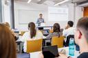Dozent steht vor einer Klasse mit mehreren Studierenden, die auf Laptops schauen, im Hintergrund eine Projektion und ein Whiteboard