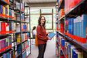 Eine junge Frau steht zwischen 2 Bücherregalen in einer Bibliothek. Sie hält ein Buch in der Hand und blickt direkt in die Kamera.