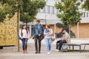 3 Studierende laufen sich unterhaltend vor einem Gebäude des TechCampus. Ein Student im Hintergrund sitzt auf eiern Bank und schaut in seinen Laptop.