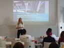 Eine Frau mit blonden Haaren steht vor einer Gruppe jungern Menschen und hält einen Vortrag zu einer Power Point Präsentation