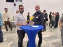 Zwei Männer an einem Stehtisch mit blauer Husse. Darauf Blumen, im Hintergrund Menschen die sich unterhalten.