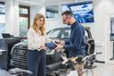 Eine junge Frau und ein junger Mann stellen etwas an einer VR-Brille ein. Im Hintergrund ist die Karosserie eines Fahrzeugs (AUDI) zu sehen.