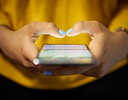 Nahaufnahme von zwei Händen, die ein Smartphone bedienen. Die Person trägt ein gelbes Oberteil und hat hellblau lackierte Fingernägel. Der Bildschirm des Smartphones ist eingeschaltet und zeigt Aktivität.