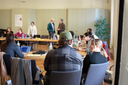 Gruppe von Menschen sitzt an einem langen Tisch in einem Besprechungsraum, zwei Personen stehen im Hintergrund und unterhalten sich.