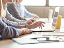 4 Personen sitzen in einer Reihe an einem Tisch mit Laptops vor sich.
